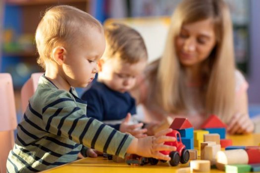 Zwei Kleinkinder spielen mit Baukl&ouml;tzen. Im Hintergrund ist eine Erzieherin zu sehen. 