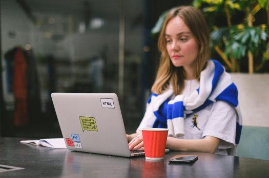 Eine Frau sitzt vor einem Laptop.
Auf dem Laptop sind Aufkleber.
Der Laptop steht auf einem Tisch, daneben ist ein Kaffeebecher.