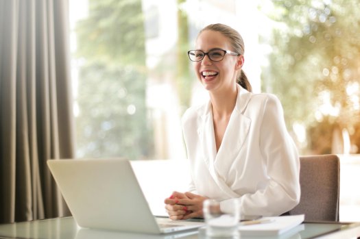 Eine junge Frau lacht. Sie trägt eine Brille.
Sie sitzt vor einem Laptop an einem Schreibtisch.
