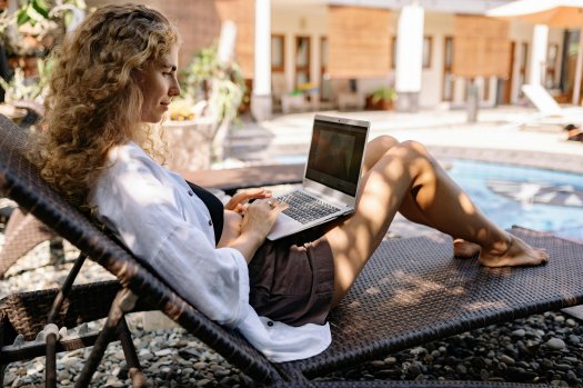 Eine Frau sitzt auf einer Liege an einem Pool.
Sie arbeitet an einem Laptop.
